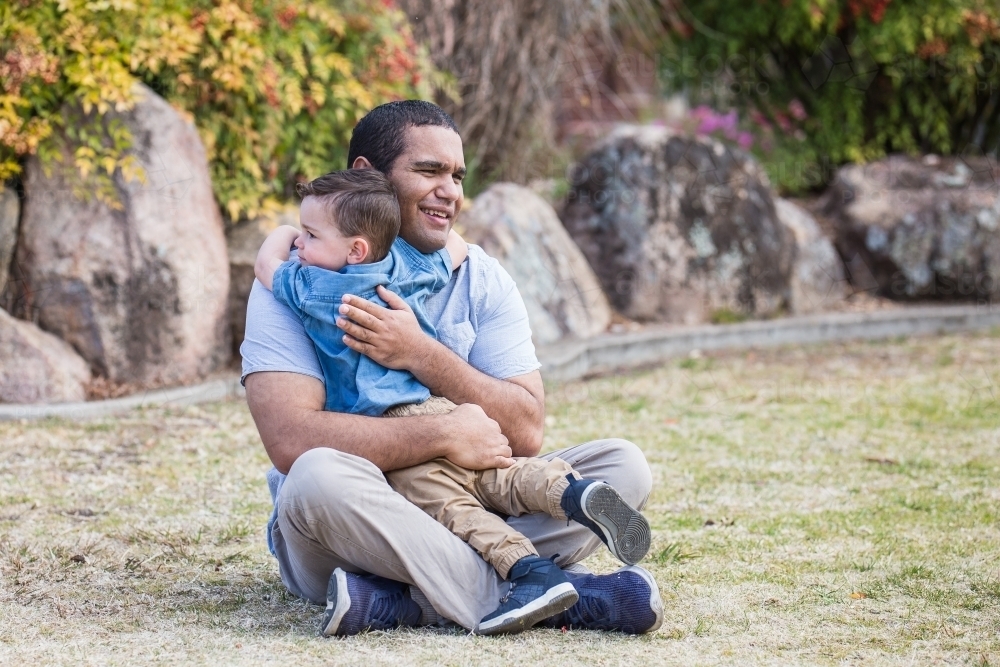 Aboriginal father sitting on grass hugging mixed race aboriginal caucasian son - Australian Stock Image