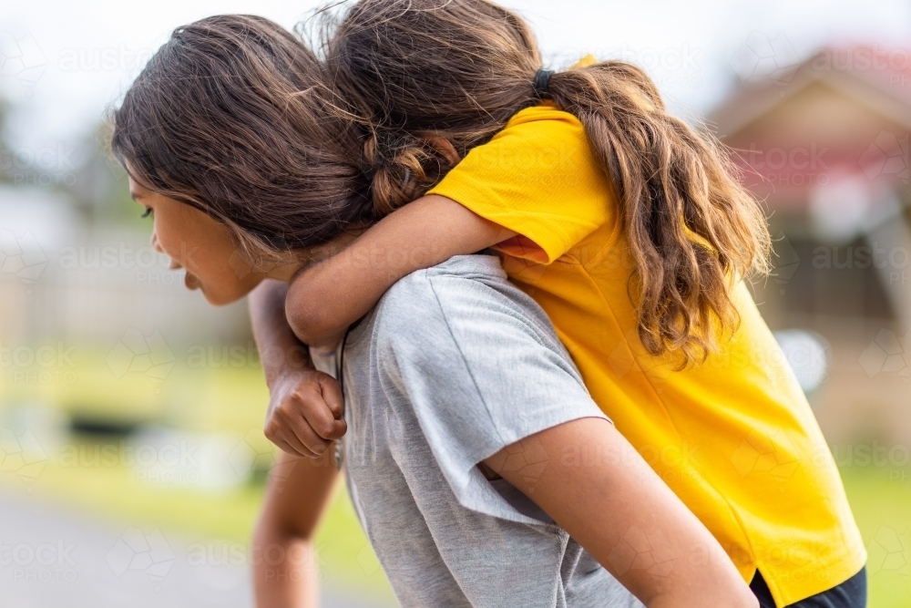 aboriginal girl piggy-backing her younger cousin with blurry background - Australian Stock Image