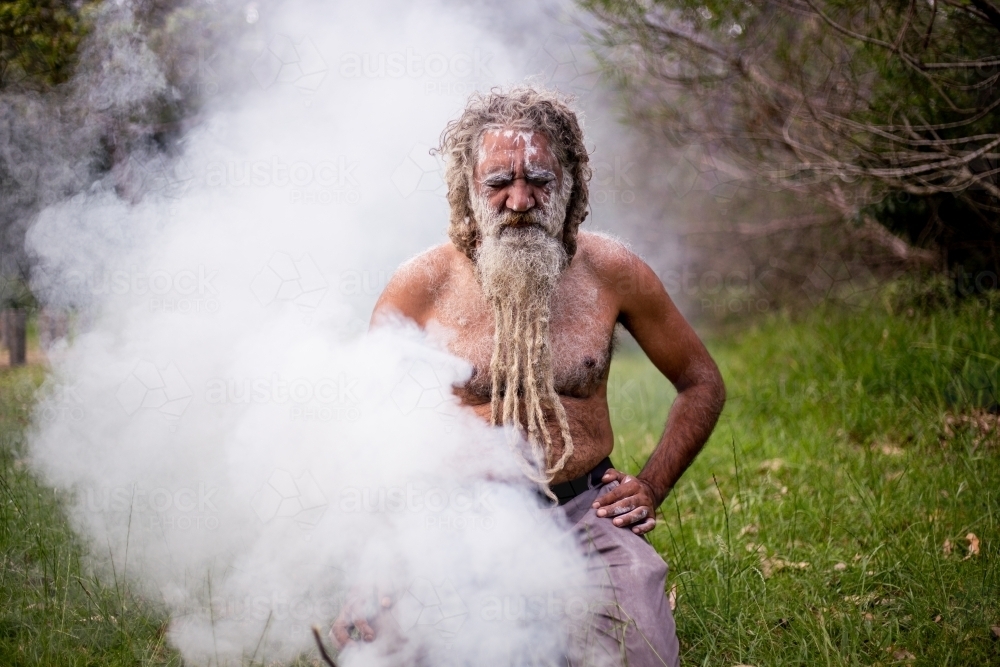 Aboriginal man kneeling behind smoke at a smoking ceremony - Australian Stock Image