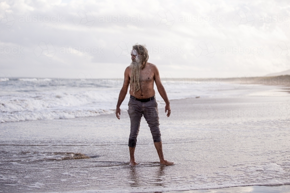 Aboriginal man standing in ankle-deep water on a beach - Australian Stock Image