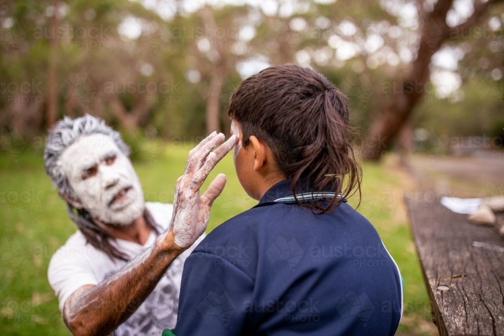 Aboriginal man wearing white face paint putting white face paint onto a young Aboriginal boy - Australian Stock Image