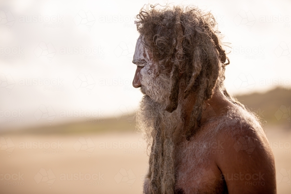 Aboriginal middle aged man with clay face paint standing on a beach looking into the distance - Australian Stock Image