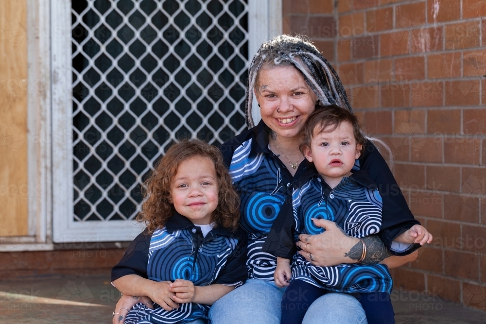 Aboriginal mum with two young daughters sitting together on front doorstep of home - Australian Stock Image