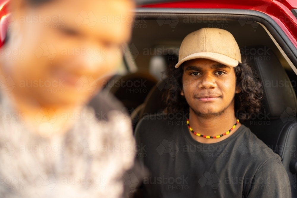 aboriginal teenaged boy sitting in car looking at camera with blurred second person in foreground - Australian Stock Image