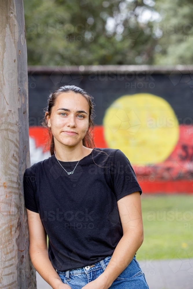 Aboriginal woman leaning against a gumtree in front of an Aboriginal flag mural in a park - Australian Stock Image