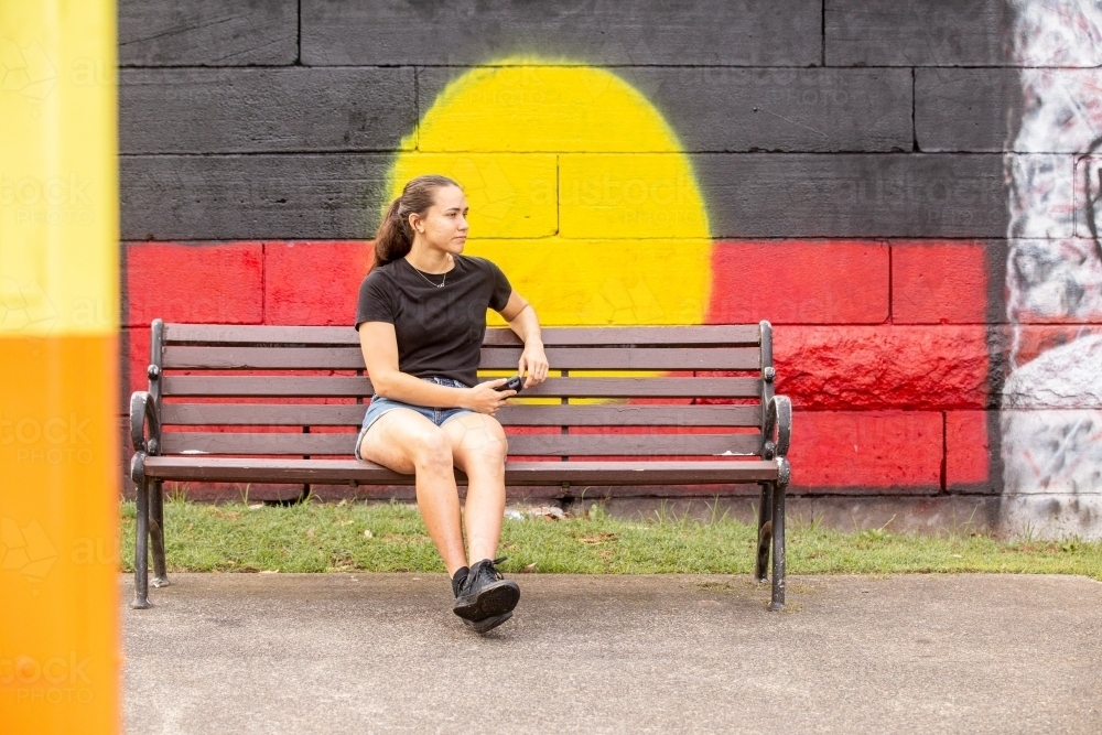 Aboriginal woman sitting on a park bench in front of a mural painting of a flag - Australian Stock Image