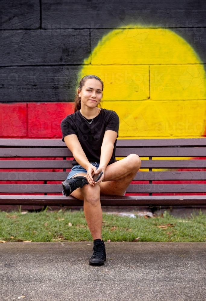 Aboriginal woman sitting on a park bench in front of a mural painting of a flag - Australian Stock Image