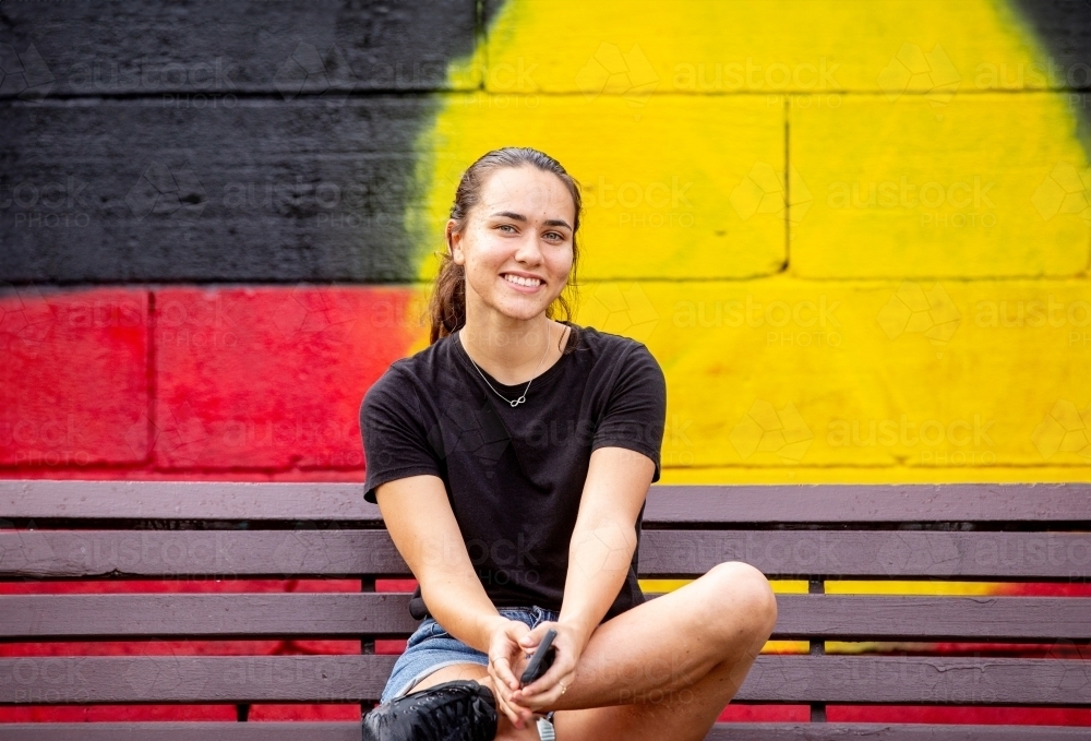 Aboriginal woman sitting on a park bench in front of a mural painting of a flag - Australian Stock Image