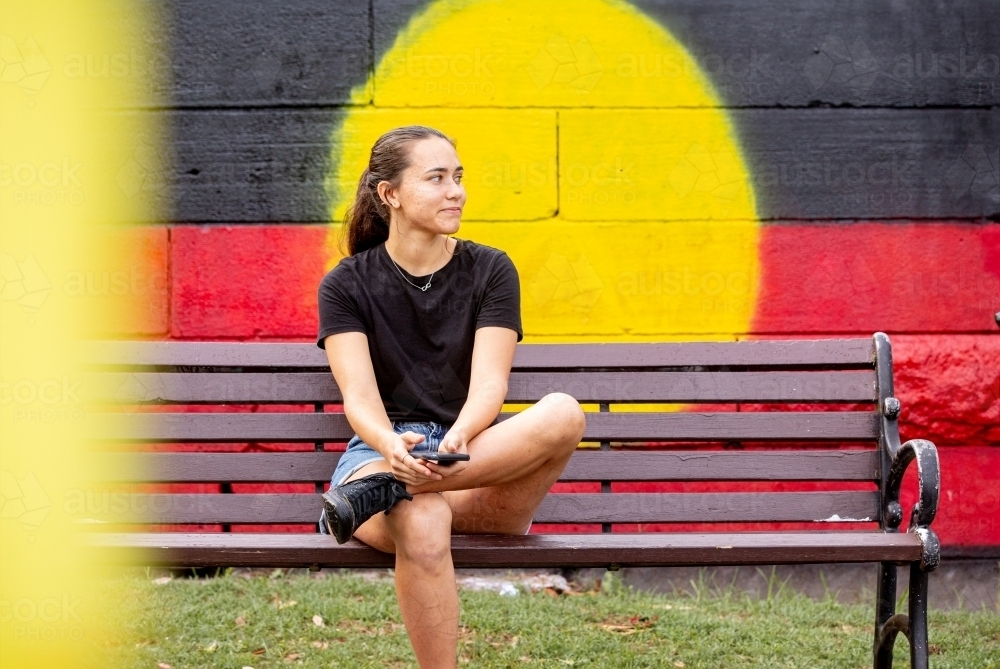 Aboriginal woman sitting on a park bench in front of a mural painting of a flag - Australian Stock Image