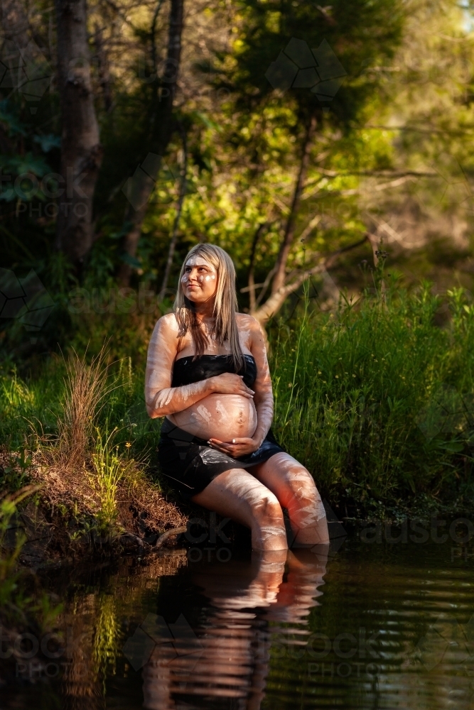 Aboriginal woman sitting on the edge of the creek with rippling sunlight on skin - Australian Stock Image