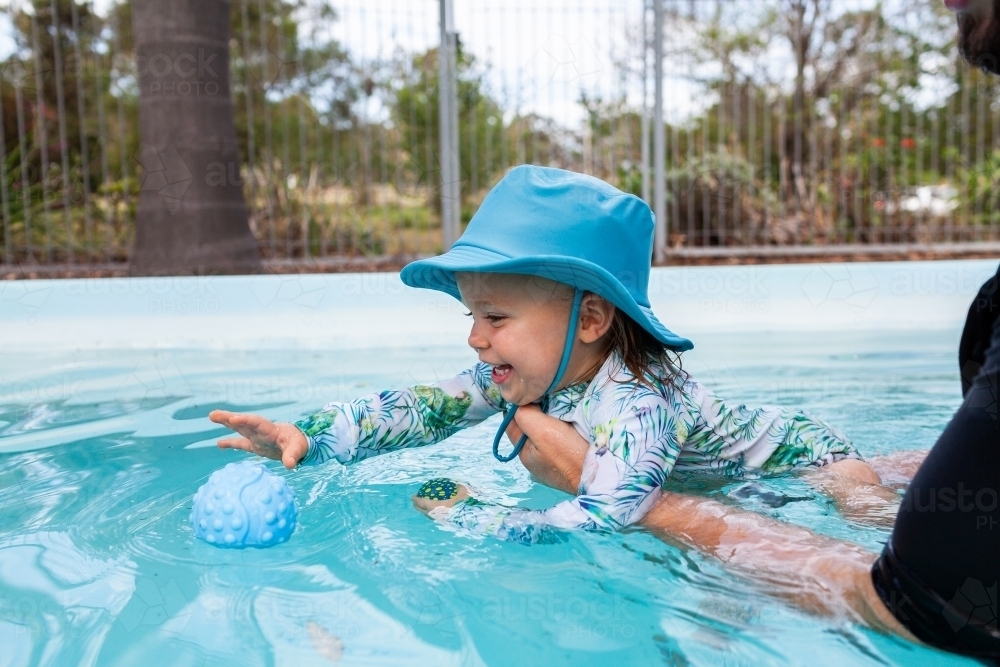 Adult holding toddler kid in backyard swimming pool practicing kicking - Australian Stock Image