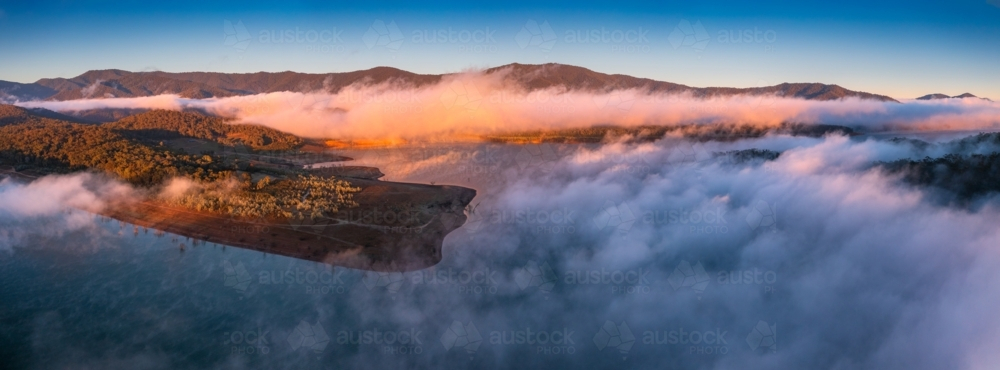 Aerial panorama of early morning light on clouds of fog and hills surrounding a high country lake - Australian Stock Image