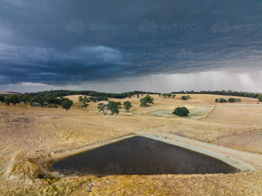 Aerial view a triangular dam on rural farmland with rain falling dark clouds above dark - Australian Stock Image