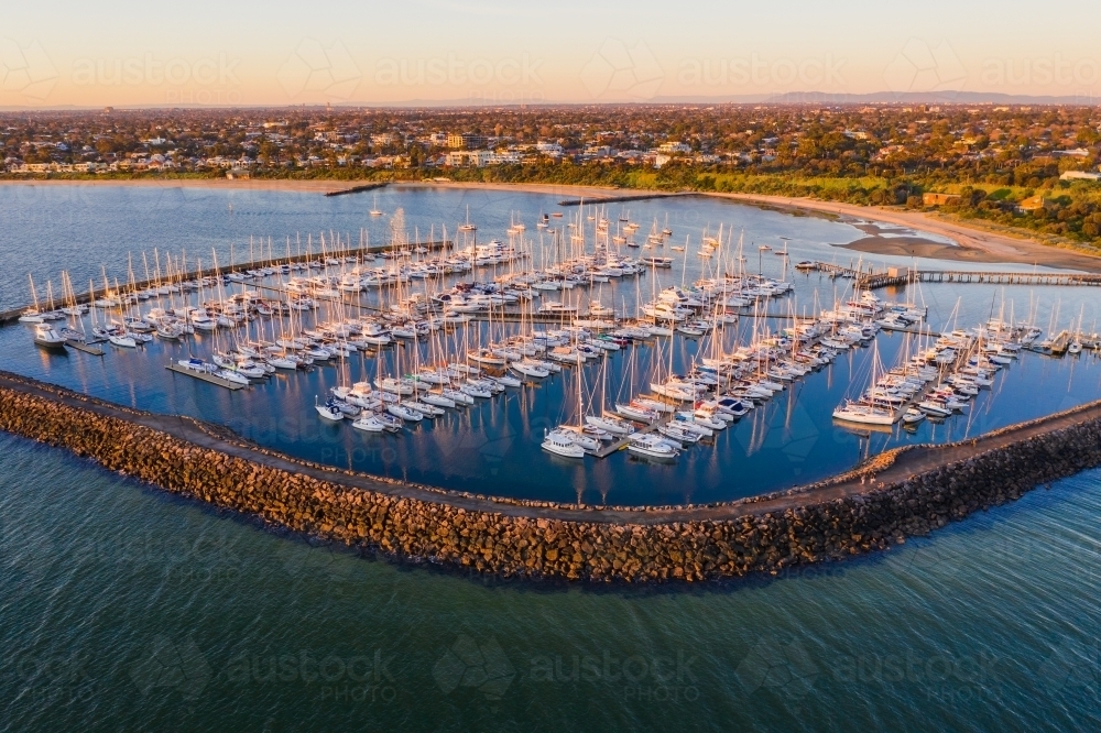 Aerial view of a bay side marina with rows of boats tied to jetties behind a large rock breakwater - Australian Stock Image