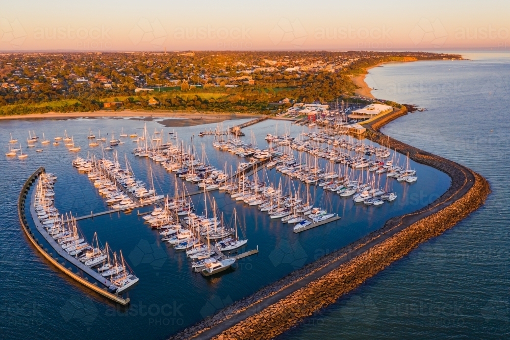 Aerial view of a bay side marina with rows of boats tied to jetties behind a large rock breakwater - Australian Stock Image