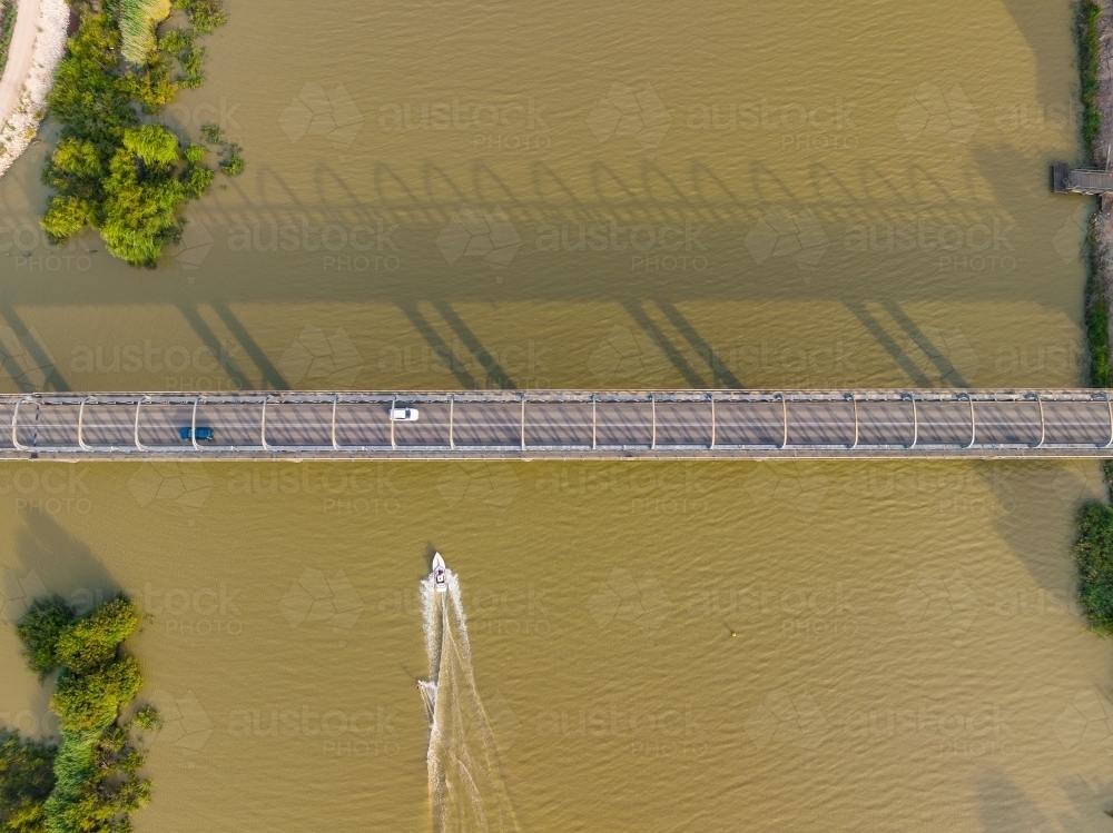 Aerial view of a boat going under a steel bridge crossing a wide brown river - Australian Stock Image