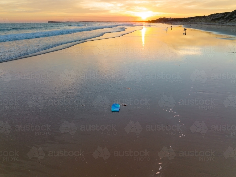 Aerial view of a boogie board and gentle waves on a beach with reflection of a sunset - Australian Stock Image