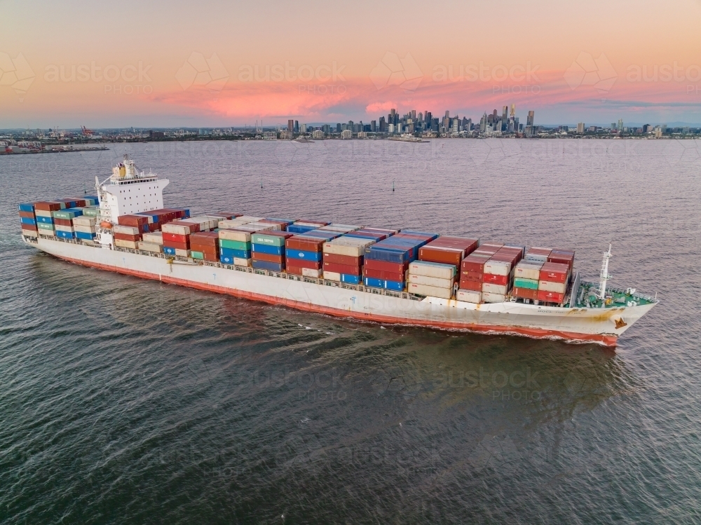 Aerial view of a cargo ship leaving port at sunset, fully laden with containers - Australian Stock Image