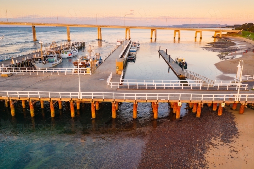 Aerial view of a coastal marina with fishing boats docked at jettys and a bridge in the background - Australian Stock Image