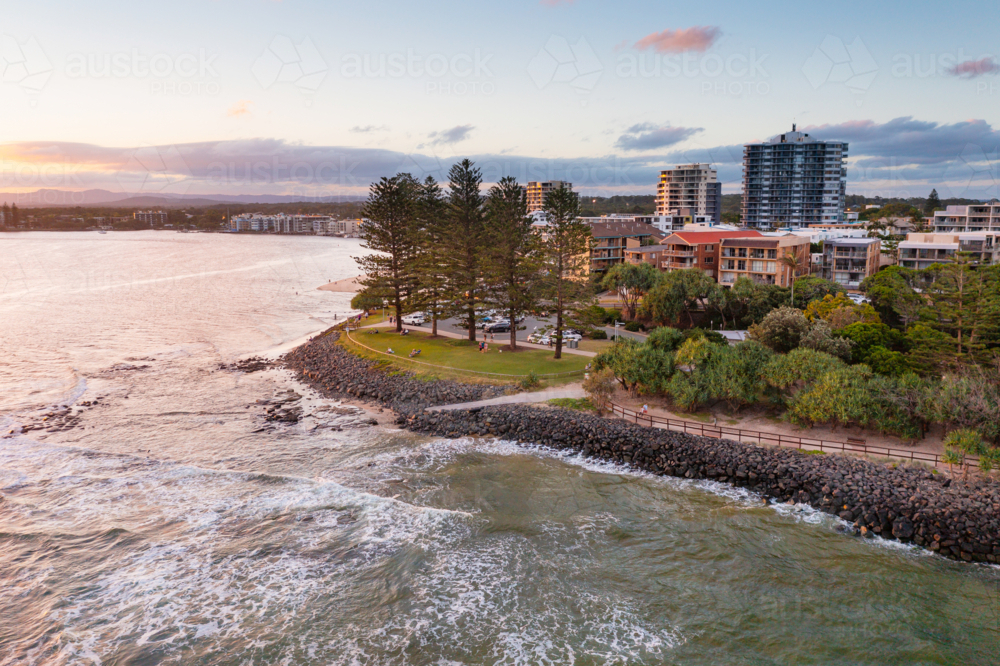 Aerial view of a coastal park with tall pine trees above a rocky breakwall at sunset - Australian Stock Image