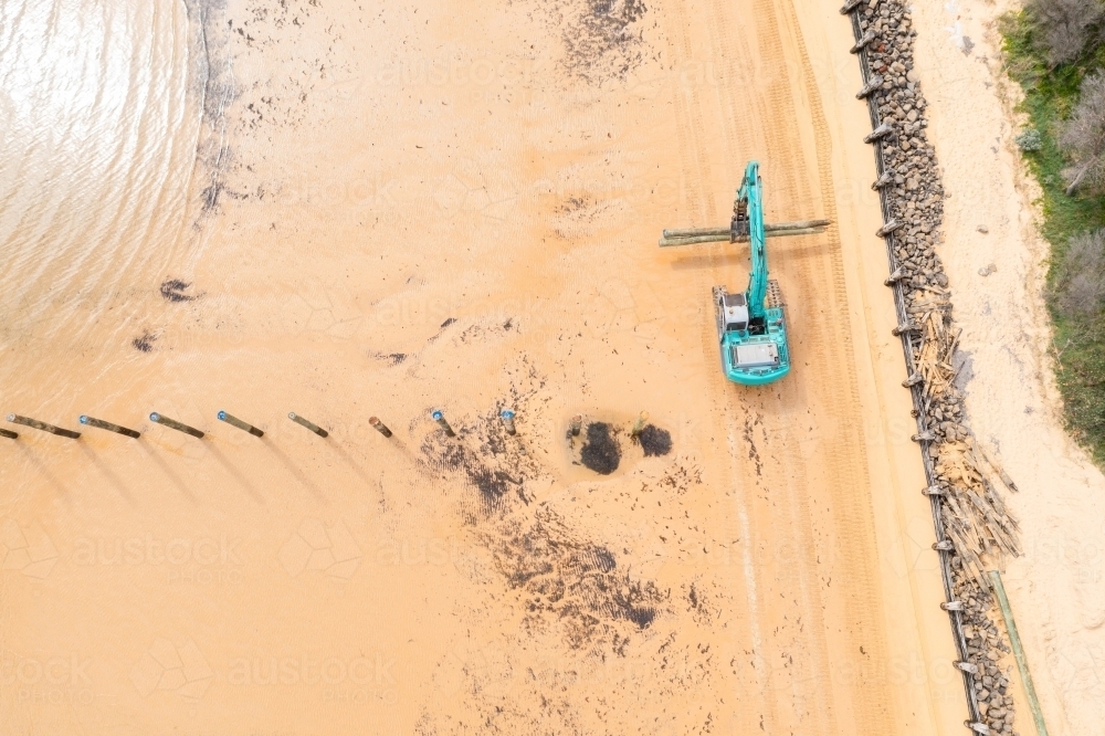 Aerial view of a colourful digger carrying poles along a sandy beach - Australian Stock Image