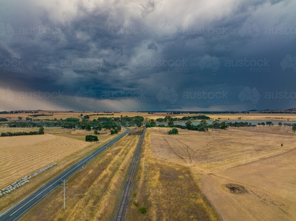 Aerial view of a country road and railway lines across dry under rain falling from a stormy sky - Australian Stock Image