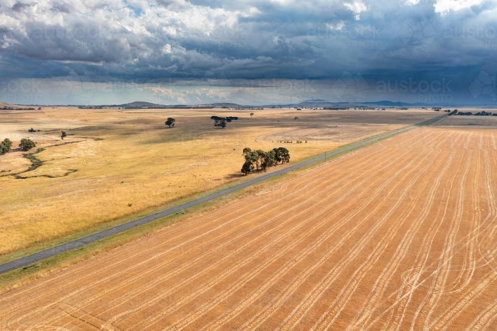 Aerial view of a country road running though dry farm land toward a rain falling from dark clouds - Australian Stock Image