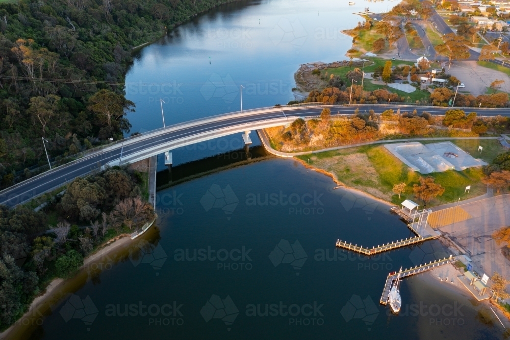 Aerial view of a curved road bridge over a river joining a forest area and a coastal town - Australian Stock Image