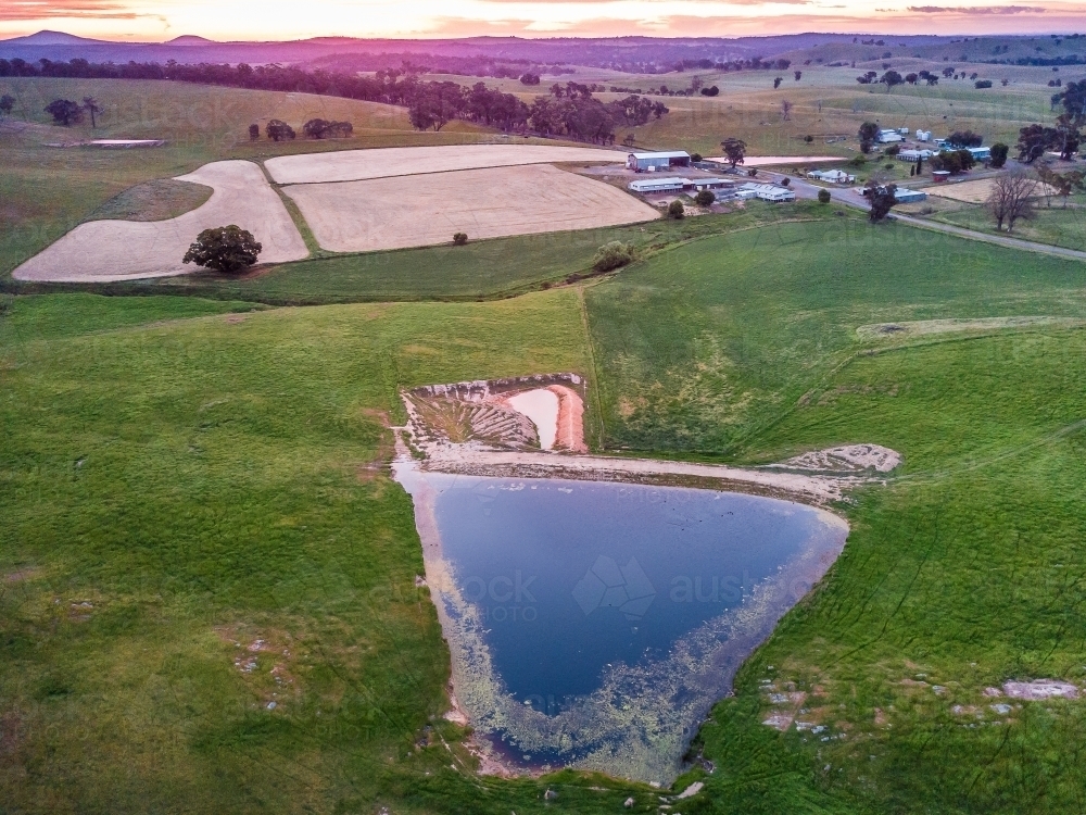 Aerial view of a dam and ploughed paddocks near farm buildings at sunset - Australian Stock Image