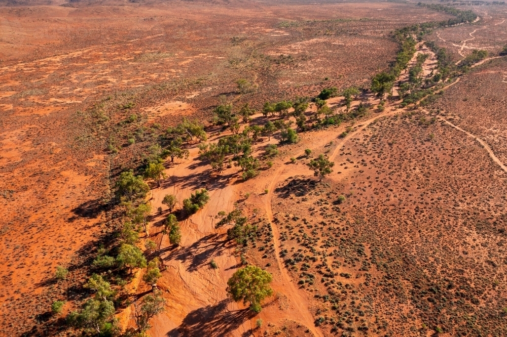 Aerial view of a dry red soil river bed lined with gum trees winding through an arid landscape - Australian Stock Image