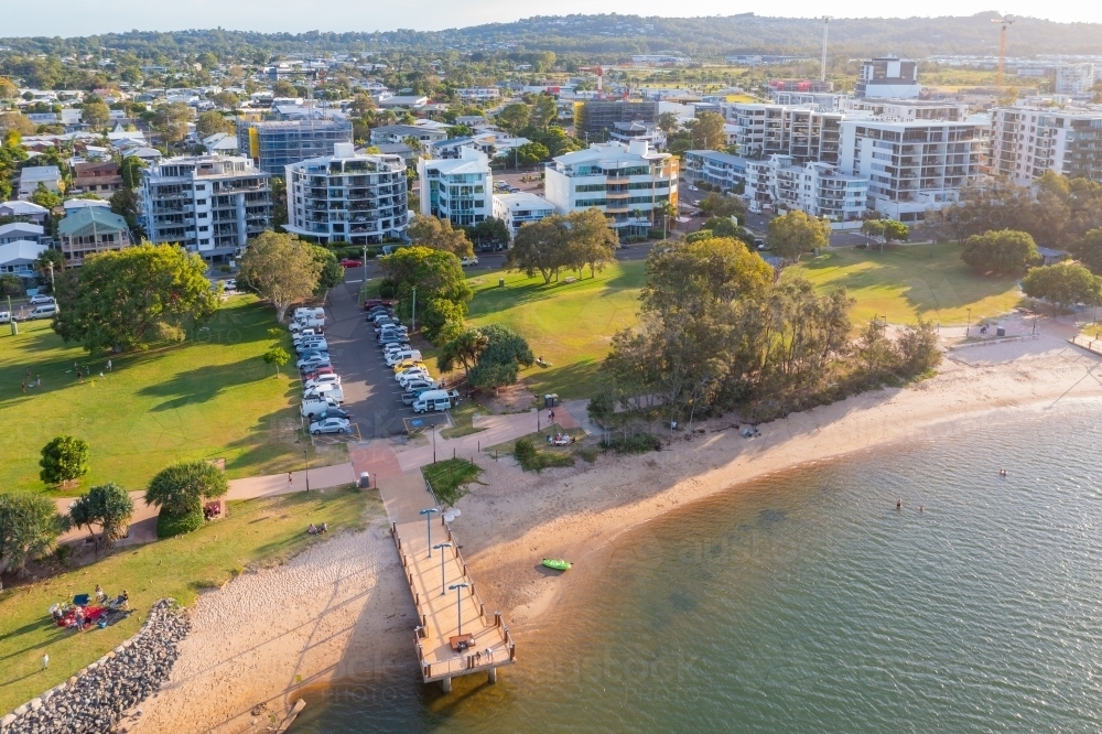 Aerial view of a jetty over a beach in front of a coastal park with high rise buildings behind - Australian Stock Image