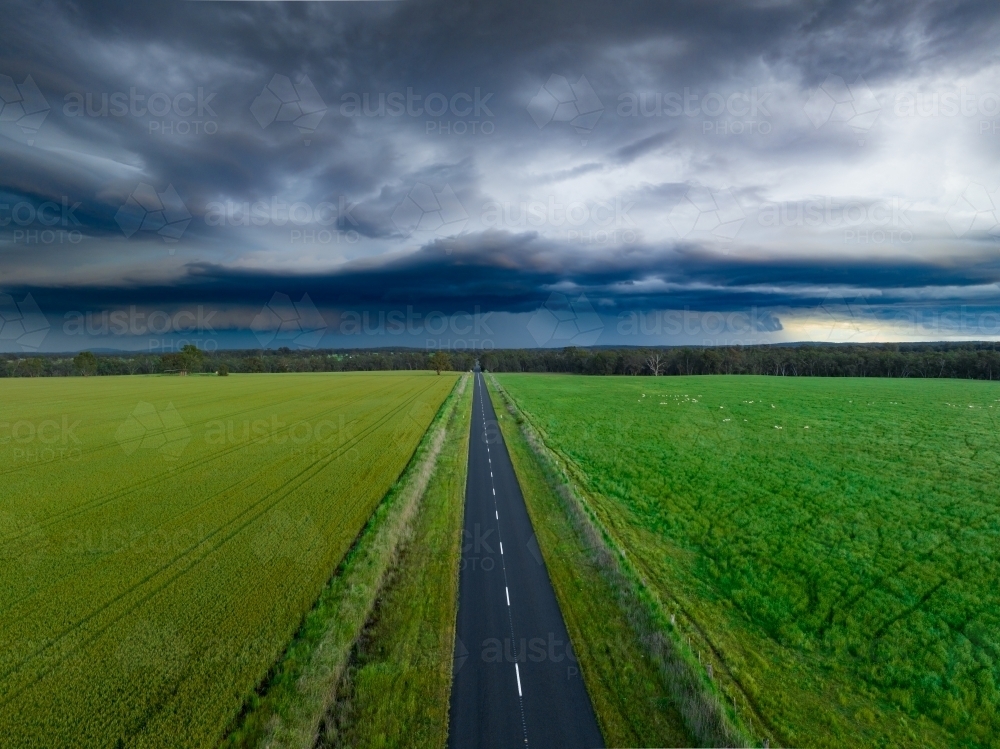 Aerial view of a narrow country road running between green farmland and under a dark stormfront - Australian Stock Image