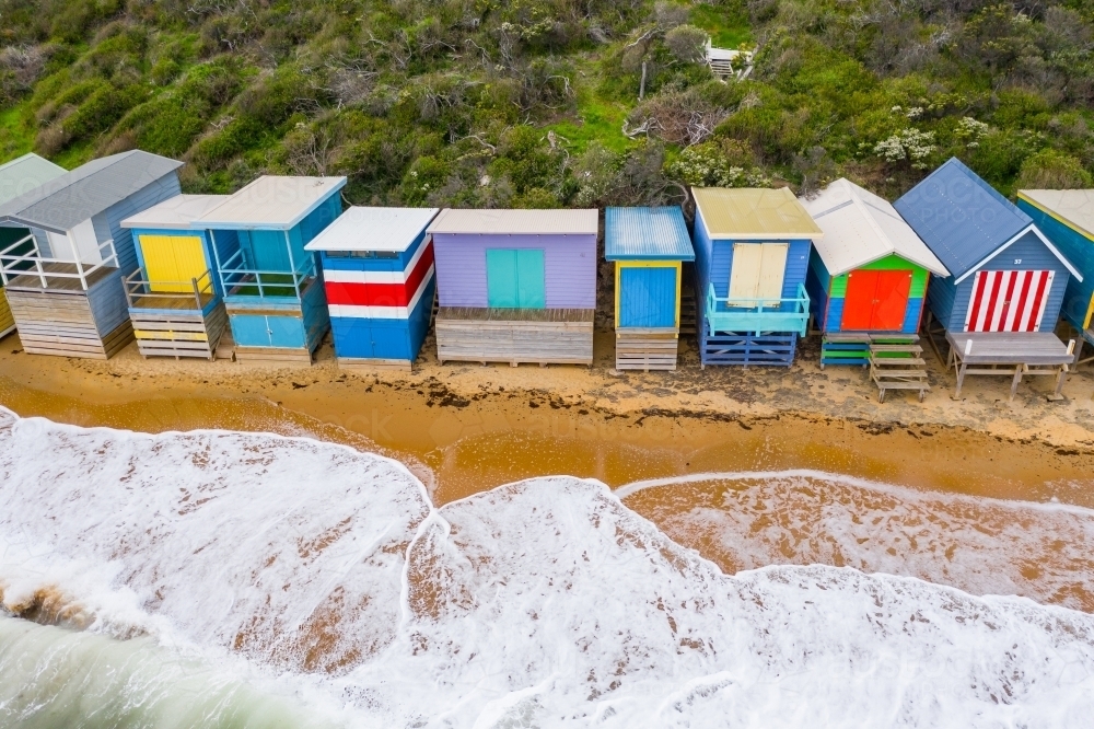 Aerial view of a row of colourful beach huts with waves washing up underneath - Australian Stock Image