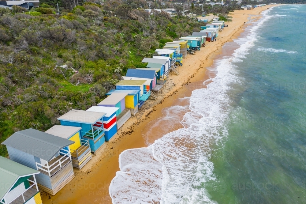 Aerial view of a row of colourful beach huts with waves washing up underneath - Australian Stock Image