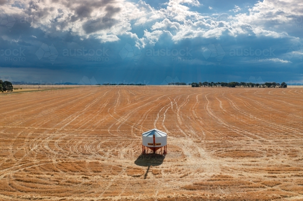 Aerial view of a solitary field bin sitting in a large barren paddock under a dark stormy sky - Australian Stock Image