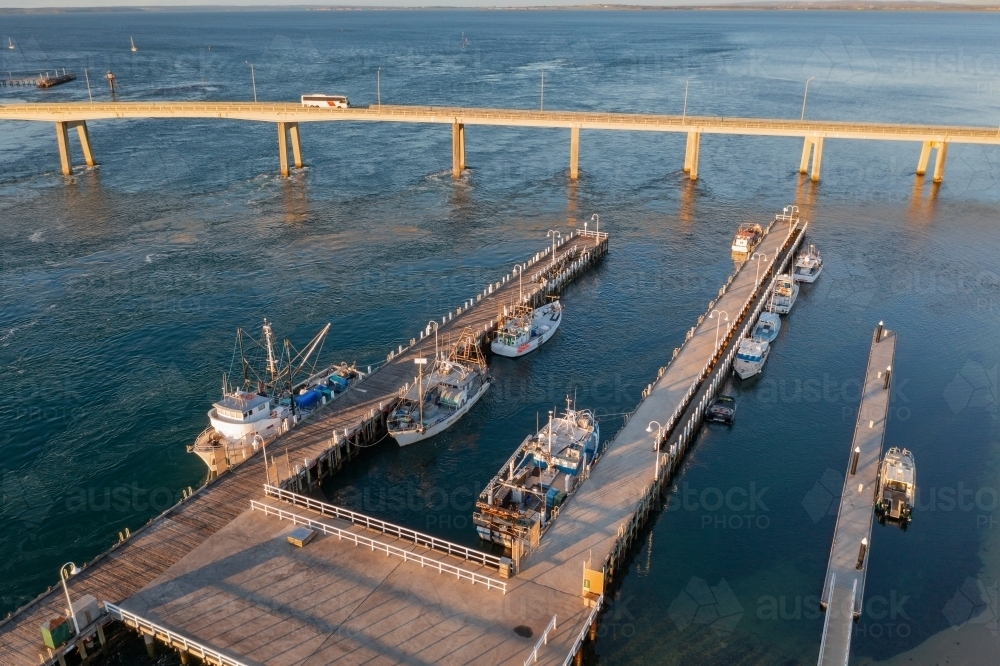 Aerial view of fishing boats docked alongside jettys near a bridge crossing a coastal bay - Australian Stock Image