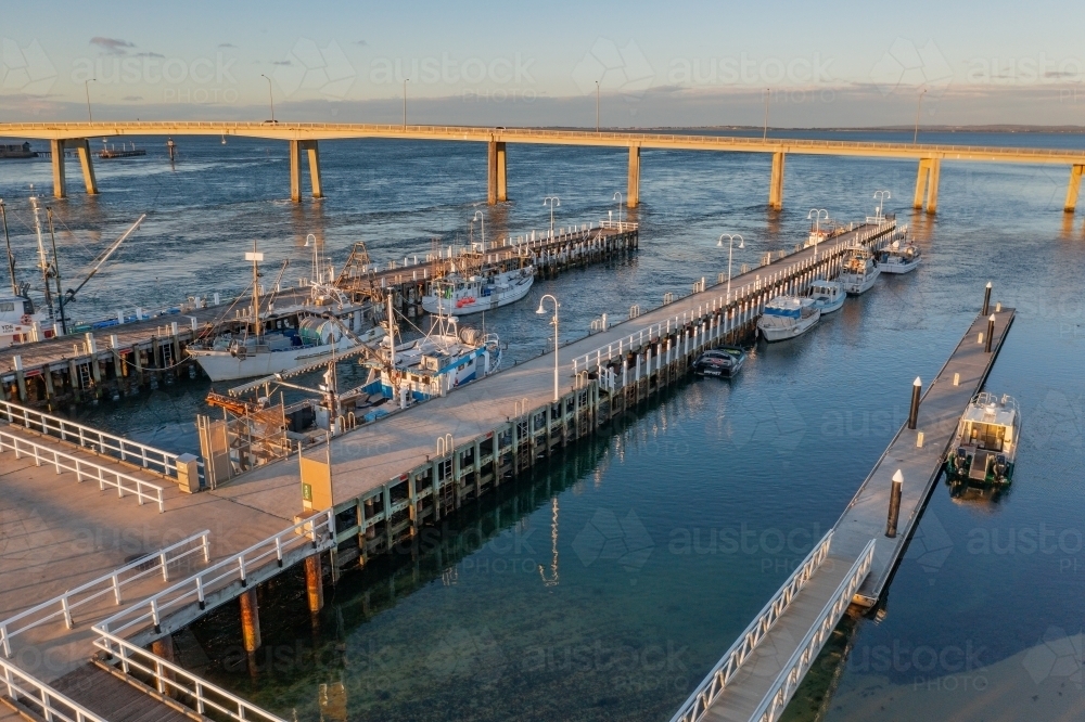 Aerial view of fishing boats docked alongside jettys near a bridge crossing a coastal bay - Australian Stock Image