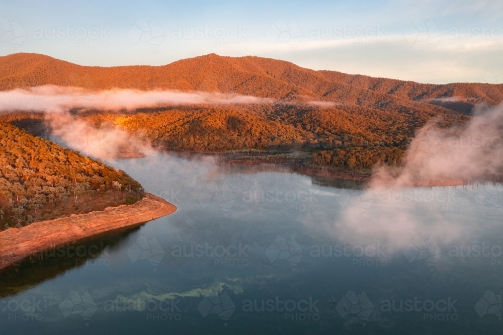 Aerial view of fog patches lifting from a lake surrounded by hills in golden morning sunlight - Australian Stock Image