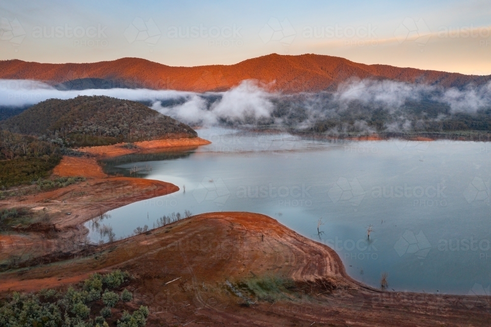 Aerial view of fog patches lifting from a lake surrounded by hills in golden morning sunlight - Australian Stock Image