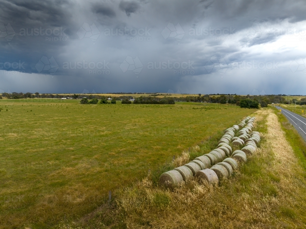 Aerial view of hay bales alongside a farm paddock with rain falling from a distant storm cloud - Australian Stock Image