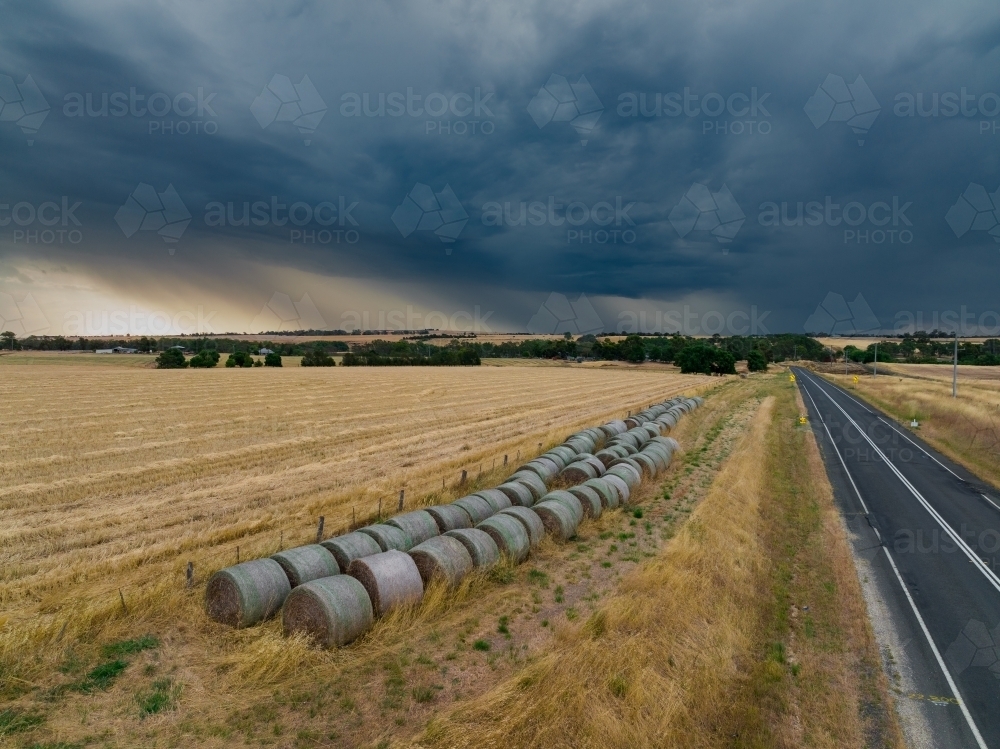 Aerial view of hay bales beside a dry paddock under rain falling from a dark stormy sky - Australian Stock Image