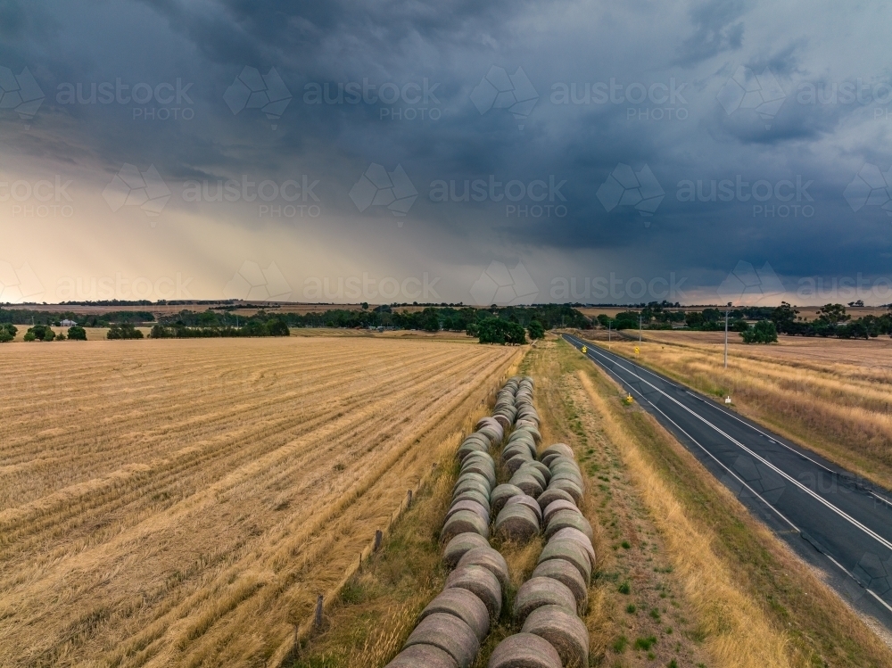 Aerial view of hay bales beside a dry paddock under rain falling from a dark stormy sky - Australian Stock Image