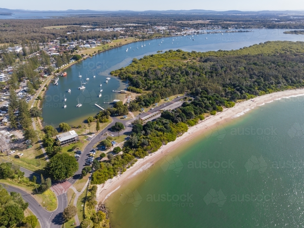 Aerial view of narrow sandy beach in front of coastal vegetation and a curved bay full of boats - Australian Stock Image