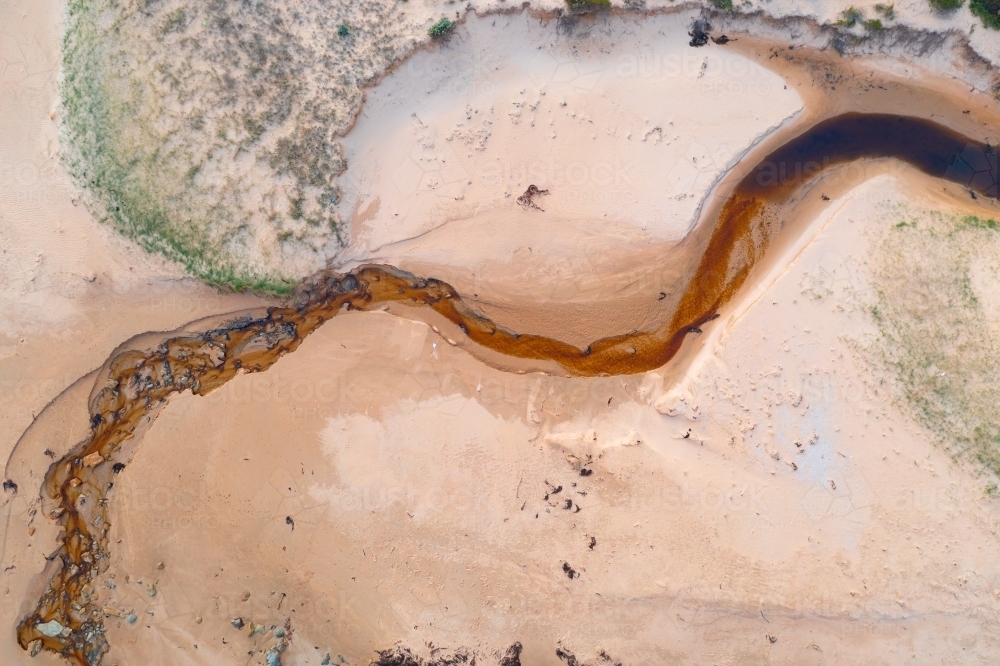 Aerial view of tannin stained creek winding its way over a sandy beach - Australian Stock Image