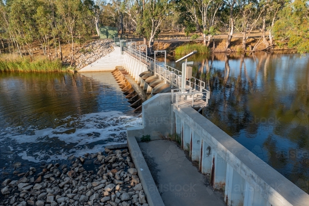 Aerial view of water flowing over a weir on a calm river with gum trees on the banks - Australian Stock Image