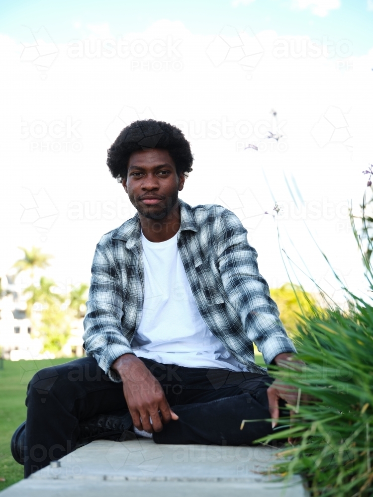 African american man sitting on a concrete bench wearing a checkered polo shirt - Australian Stock Image