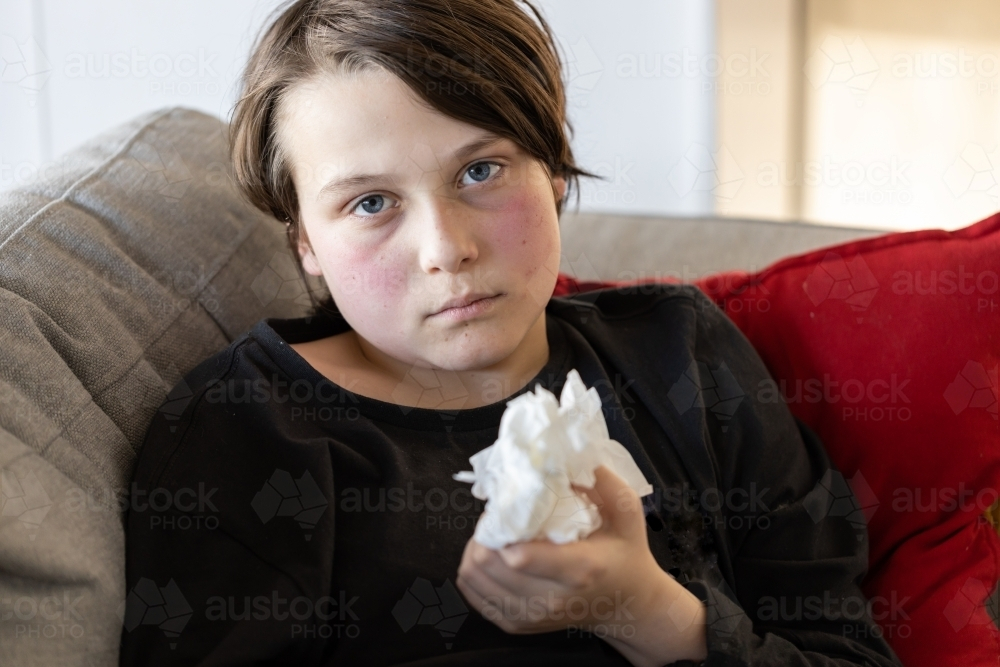 An unwell sick preteen boy lying on a couch holding some used tissues - Australian Stock Image