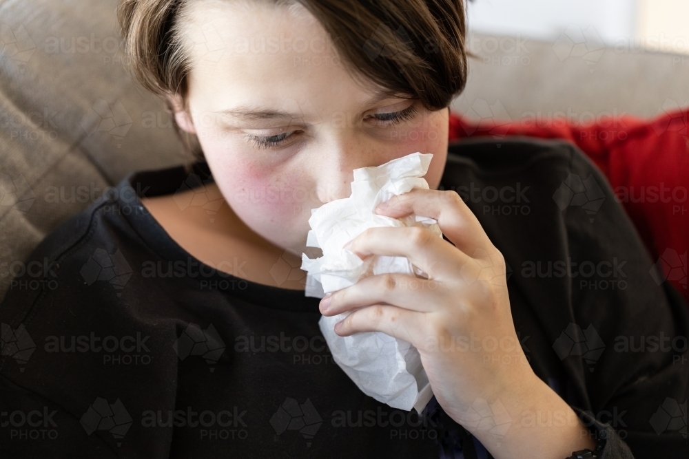 An unwell sick preteen boy lying on a couch holding tissues to his nose, his cheeks are red - Australian Stock Image