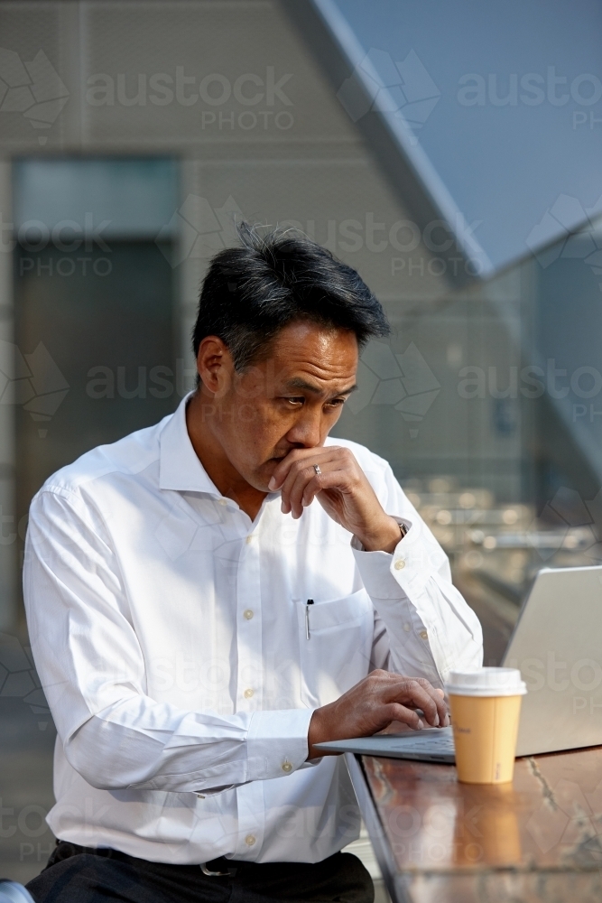 Asian businessman having a coffee whilst using laptop - Australian Stock Image