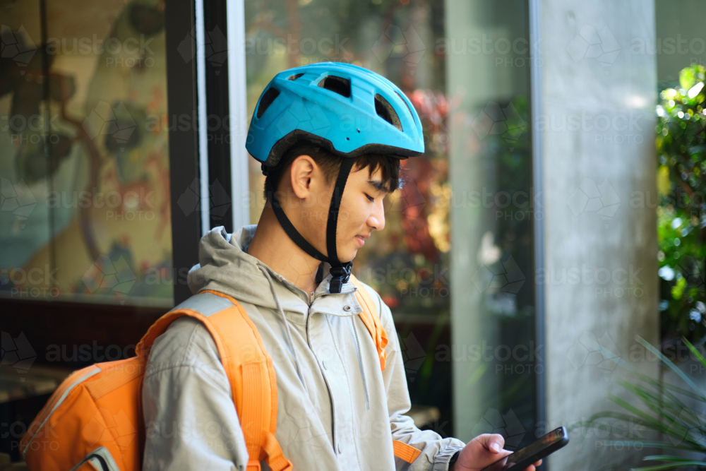Asian delivery guy looking on his phone outside restaurant wearing bike helmet - Australian Stock Image